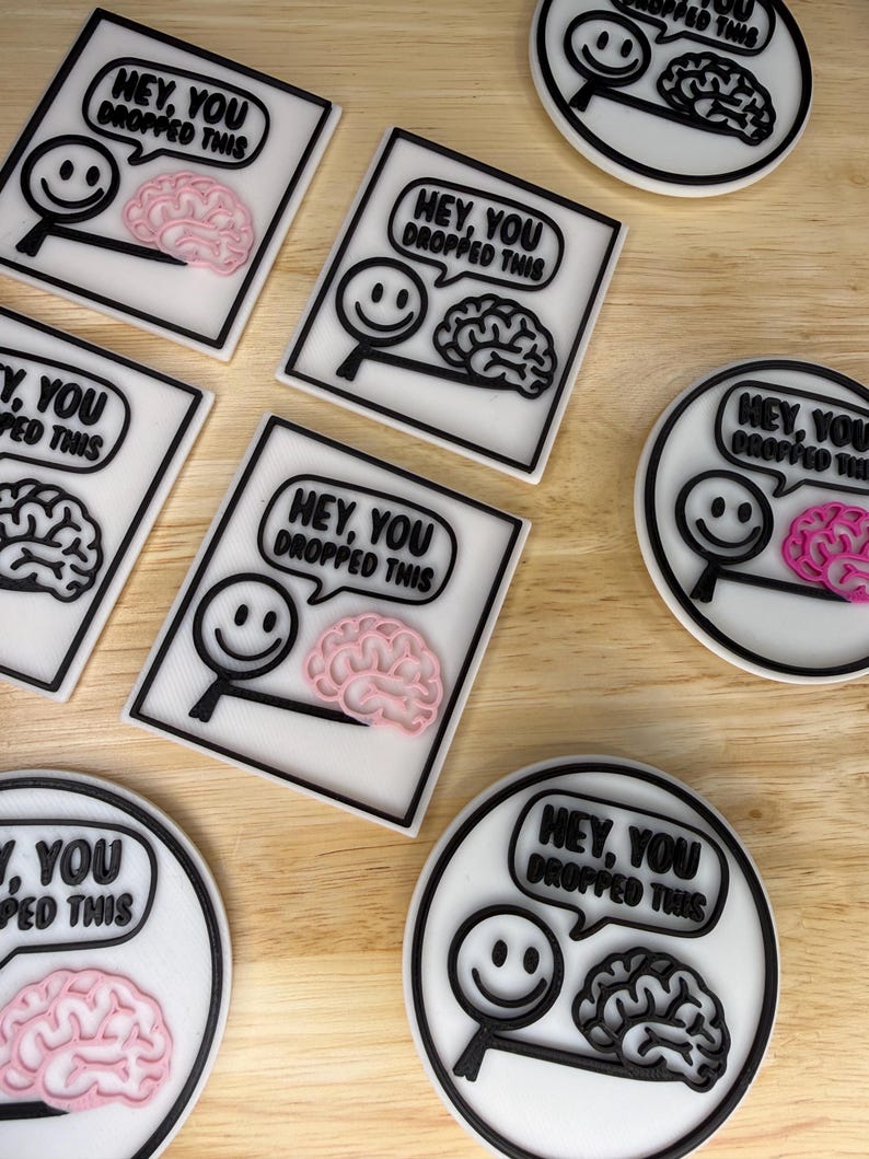 May include: Coasters with a black and white design featuring a smiley face, magnifying glass, and brain graphic. The text "HEY, YOU DROPPED THIS" is in a speech bubble. Some coasters have a pink brain, others a black brain.
