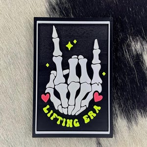 May include: Black rectangular sign with a white border featuring a skeleton hand making a rock-and-roll gesture. The words "LIFTING ERA" are in neon green, with two pink hearts and yellow stars.