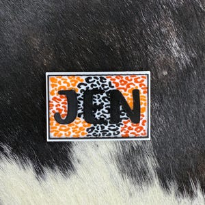 May include: A rectangular patch with the letters "JEN" in black, set against a background of orange and black leopard print. The patch is outlined in white.