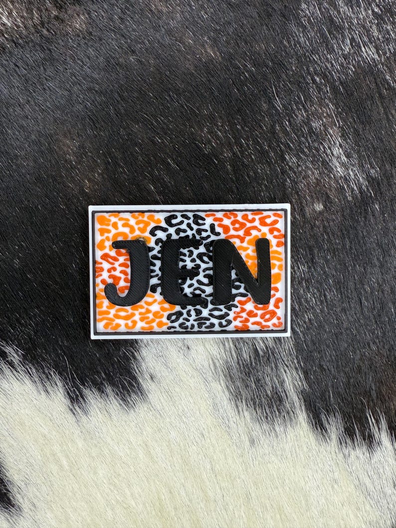 May include: A rectangular patch with the letters "JEN" in black, set against a background of orange and black leopard print. The patch is outlined in white.