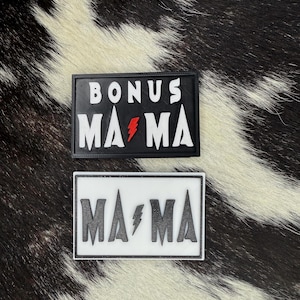 May include: Two rectangular patches. The top patch reads "BONUS MAMA" with a red lightning bolt, the bottom patch reads "MAMA" with a white lightning bolt. The text is white on a black background. The background is a black and white cowhide.