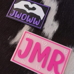 May include: Two rectangular patches. The top patch has the text "JWOWW" in purple, with a white hand-heart design on a black background. The bottom patch has the letters "JMR" in pink, with small heart designs on a pink background.