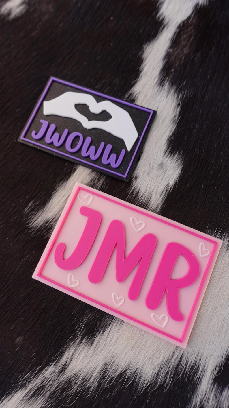 May include: Two rectangular patches. The top patch has the text "JWOWW" in purple, with a white hand-heart design on a black background. The bottom patch has the letters "JMR" in pink, with small heart designs on a pink background.
