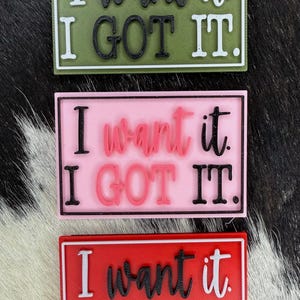 May include: Three rectangular patches with the text "I want it. I GOT IT." in black and white. The patches are in olive green, pink, and red, each with a white border. The patches are displayed on a black and white cowhide background.