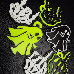 May include: A collection of Halloween-themed charms in various designs. The charms include a skeleton hand giving the middle finger, and a cartoon ghost. The charms are in black, white, and neon green colors.