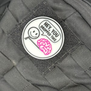 May include: A circular white patch with a black border, displaying a cartoon magnifying glass with a smiley face and a pink brain. Text in a speech bubble reads "Hey, you dropped this". The patch is attached to a black fabric.