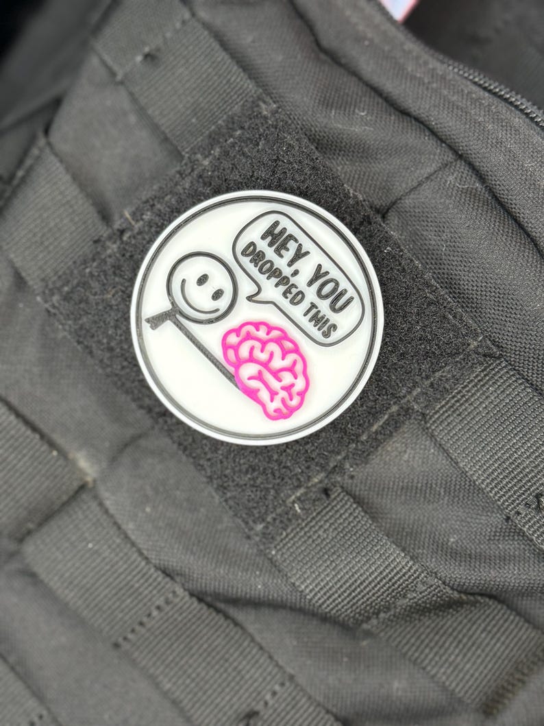 May include: A circular white patch with a black border, displaying a cartoon magnifying glass with a smiley face and a pink brain. Text in a speech bubble reads "Hey, you dropped this". The patch is attached to a black fabric.