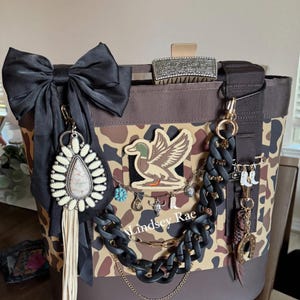 May include: A brown and tan camouflage tote bag with a black bow, decorative charms, and a duck emblem. The bag features a black chain and gold accents, with the text "Lindsey Rae".