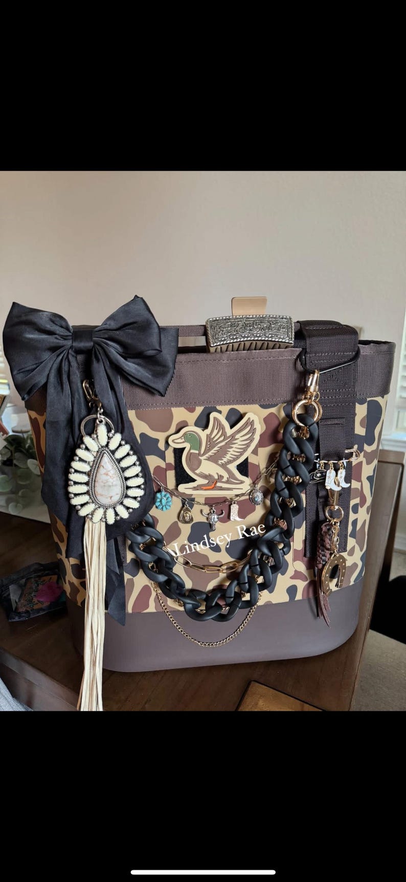May include: A brown and tan camouflage tote bag with a black bow, decorative charms, and a duck emblem. The bag features a black chain and gold accents, with the text "Lindsey Rae".