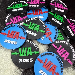 May include: A selection of circular tokens for a barbell festival. Each token displays "VEA 2025" in vibrant colours like green, pink, and red, on black and blue discs. The words "BARBELL FESTIVAL" are also printed on the tokens.