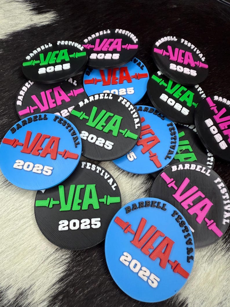 May include: A selection of circular tokens for a barbell festival. Each token displays "VEA 2025" in vibrant colours like green, pink, and red, on black and blue discs. The words "BARBELL FESTIVAL" are also printed on the tokens.