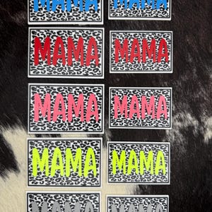 May include: A collection of ten rectangular patches featuring a black and white leopard print background. Each patch displays the word MAMA in bold, sans-serif capital letters. The text colors include blue, red, pink, neon yellow, and gray.