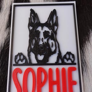 May include: A white rectangular sign with a black border features a black German Shepherd dog design and the name "SOPHIE" in red letters. The dog's paws are visible at the bottom of the design.