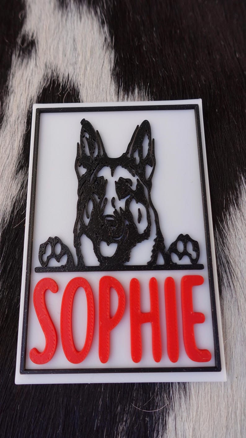 May include: A white rectangular sign with a black border features a black German Shepherd dog design and the name "SOPHIE" in red letters. The dog's paws are visible at the bottom of the design.