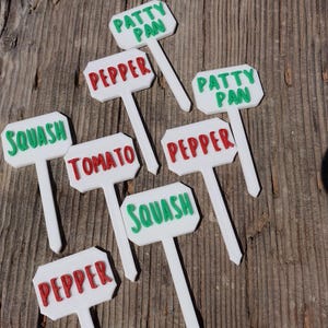 May include: White plant markers with green and red text, displaying vegetable names such as "Squash", "Pepper", "Tomato", and "Patty Pan". The markers are set on a wooden surface, ideal for a vegetable garden.