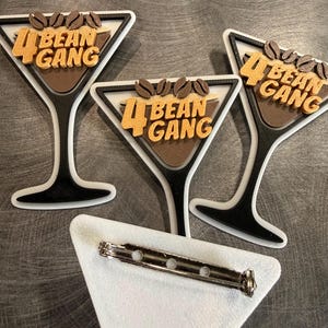 May include: Three martini-shaped pins with the text "4 Bean Gang" in orange and brown. Each pin features coffee beans at the top. The pins are white, black, and brown, with a silver pin back.