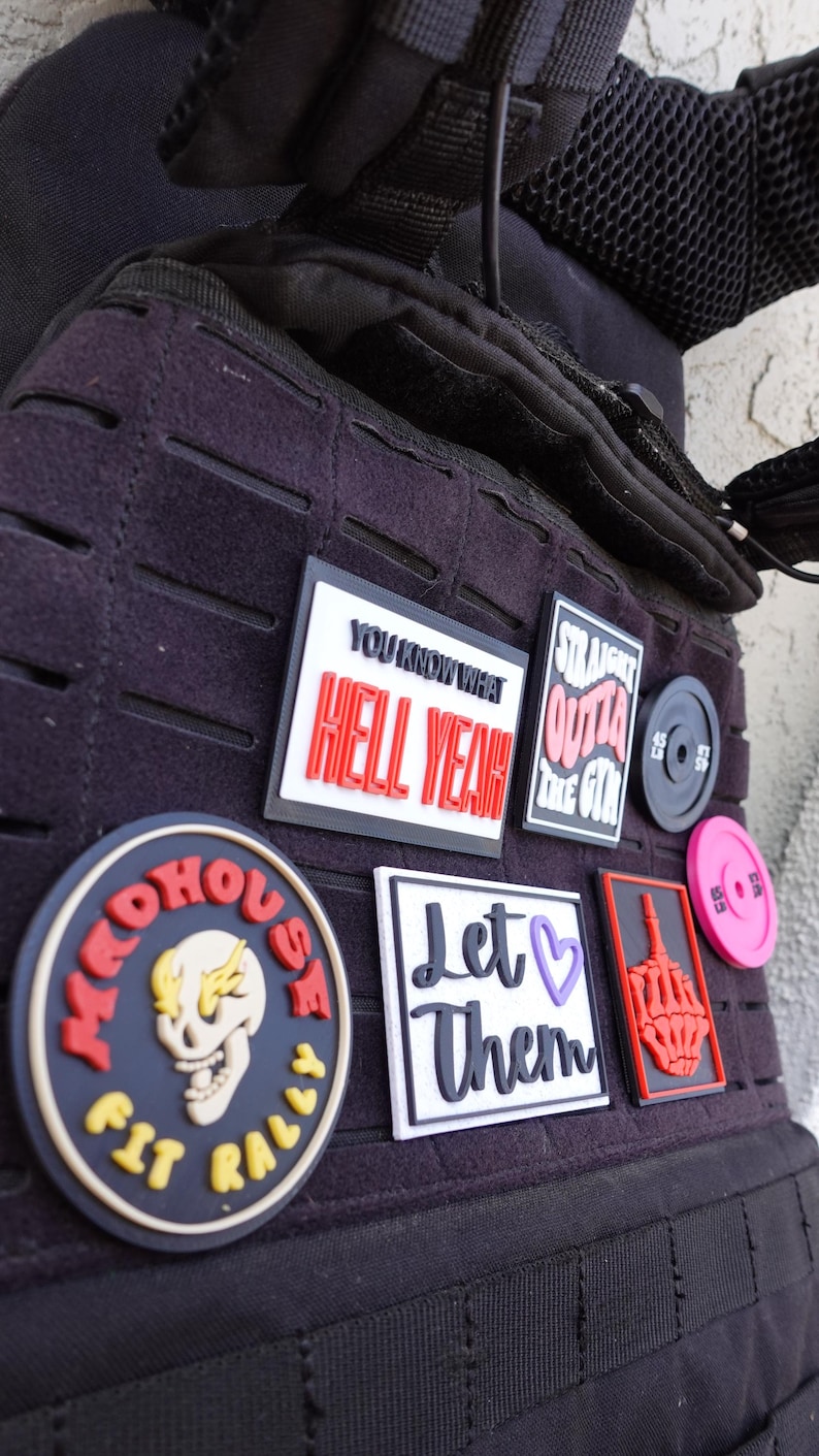 May include: A black tactical vest adorned with patches and gym-related accessories. Patches feature phrases such as "HELL YEAH" and "STRAIGHT OUTTA THE GYM." Includes a weight plate and a pink plate.