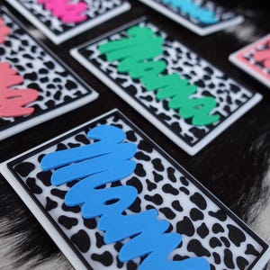 May include: Rectangular stickers with the word "Mama" in various colors, including pink, blue, and green. The stickers have a black and white cow print background and a white border. The stickers are displayed on a black and white patterned surface.