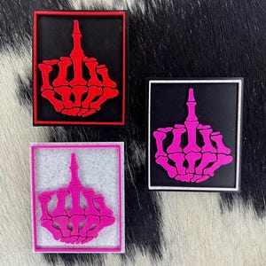 May include: Three square patches featuring a skeletal hand giving the middle finger. One patch has a red hand on a black background with a red border. Another has a pink hand with a white border. The third has a pink hand with a pink border.