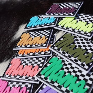 May include: A selection of 'Mama' patches in various colours. Each patch displays the word 'Mama' in shades like pink, orange, and green, set against a black and white chequered pattern. The patches are arranged on a dark, textured surface.