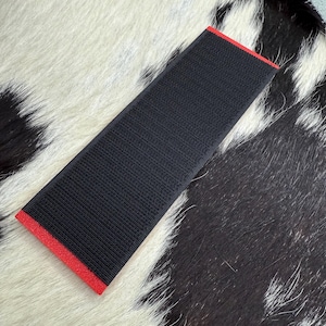 May include: A rectangular black fabric with a textured surface and a red border. The item is placed on a black and white cowhide rug.