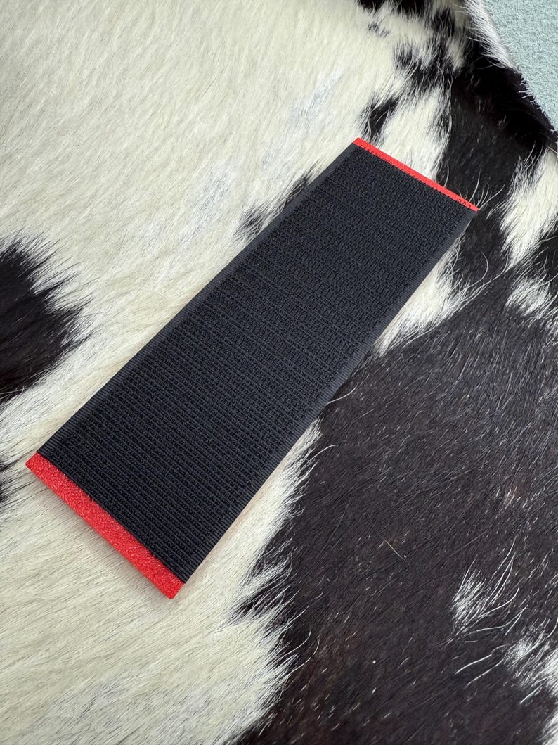 May include: A rectangular black fabric with a textured surface and a red border. The item is placed on a black and white cowhide rug.