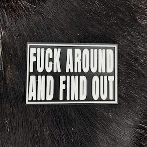 Könnte beinhalten: Ein rechteckiger schwarz-weißer Aufnäher mit dem Text "FUCK AROUND AND FIND OUT" in fetten Großbuchstaben. Der Aufnäher befindet sich vor einem dunklen, strukturierten Hintergrund.