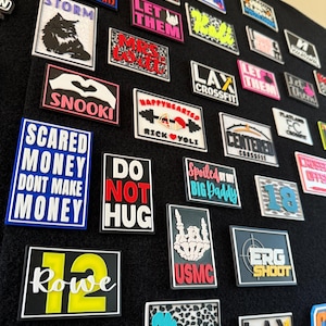 May include: A display of rectangular patches with diverse designs and text, including "Storm," "Happyhearted," and "LAX CrossFit." The patches feature a range of colours, such as blue, pink, and black, and are attached to a black surface.
