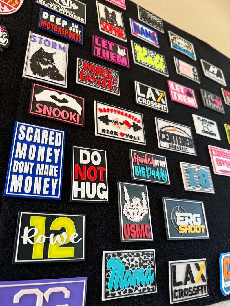 May include: A display of rectangular patches with diverse designs and text, including "Storm," "Happyhearted," and "LAX CrossFit." The patches feature a range of colours, such as blue, pink, and black, and are attached to a black surface.