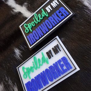 May include: Two rectangular patches with the text "Spoiled by my Ironworker". The text is in green, black, and blue, with a 3D effect. One patch has a black border, the other a white border. The background is a black and white cowhide pattern.