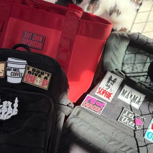 May include: A black tactical backpack with several patches, including a coffee cup, a skeleton hand, and the text "I don't give a ship." A red tote bag with the text "Shit Show Supervisor" is also visible.