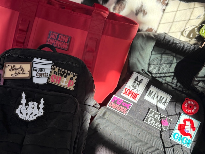 May include: A black tactical backpack with several patches, including a coffee cup, a skeleton hand, and the text "I don't give a ship." A red tote bag with the text "Shit Show Supervisor" is also visible.
