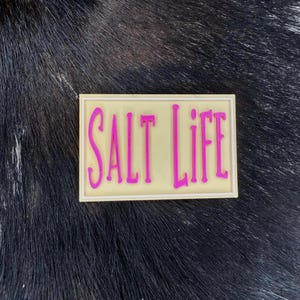 May include: A rectangular, cream-coloured sticker with the words "SALT LIFE" in bright pink, outlined in a darker pink. The sticker is set against a dark, textured background.