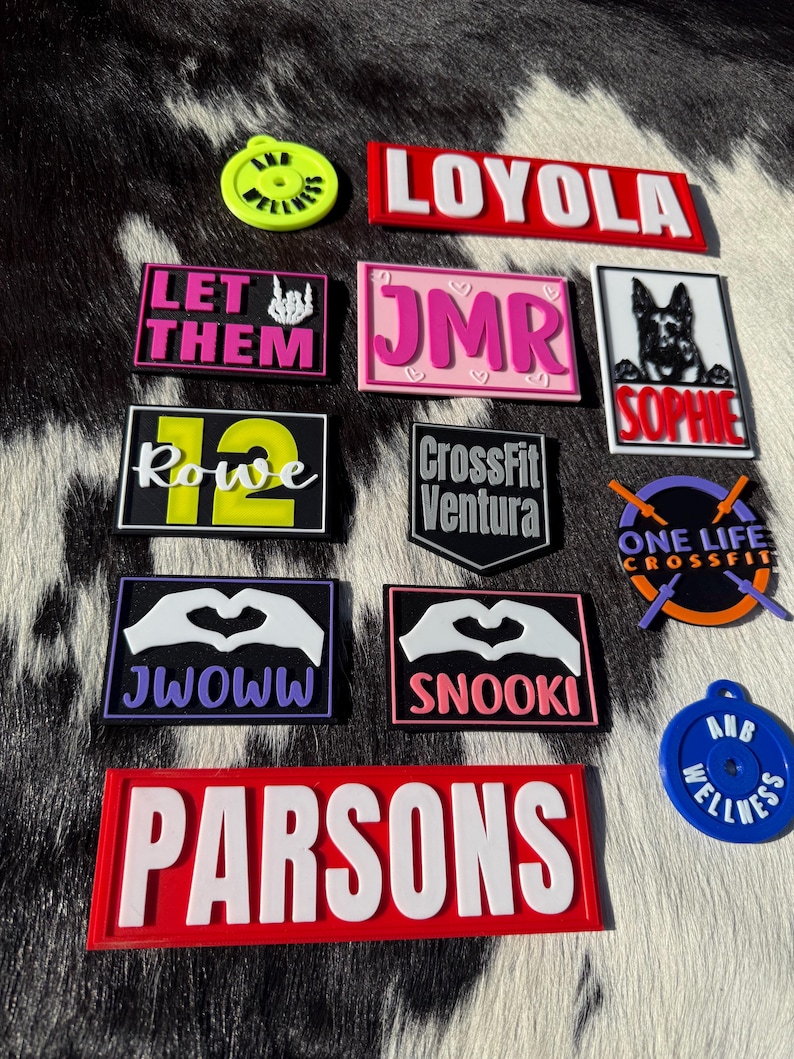 May include: A selection of vibrant, rectangular patches and circular charms, each displaying text and graphics. The patches feature names such as "LOYOLA" and "PARSONS", alongside other text and images. The charms include the text "ANB WELLNESS."