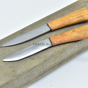 May include: Two small, sharp knives with wooden handles. The knives are laid out on a wooden surface. The blades are made of metal and have a shiny finish. The handles are made of wood and have a natural finish.