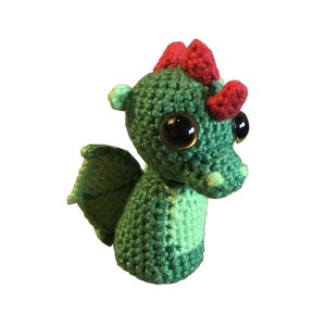 Peut inclure: Un dragon en crochet vert avec des cornes rouges et de grands yeux bruns. Le dragon a une petite aile sur le dos.