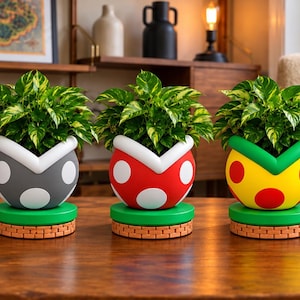 May include: Five round planters shaped like video game piranha plant pipes, each holding a variegated green and yellow pothos plant. The planters feature white or colored spots and sit on green circular bases with a brick-patterned bottom edge.