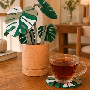 May include: A set of monstera leaf-shaped coasters in green, white, and olive, stored in a cylindrical peach-colored pot with embossed leaf patterns. A clear glass cup filled with dark tea rests on one of the coasters on a wooden table.