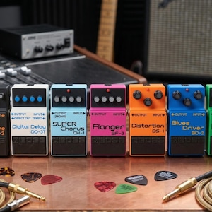 May include: A collection of seven BOSS guitar effects pedals in various colors, including black, white, blue, pink, orange, and green. Each pedal has a different name, such as Metal Zone, Digital Delay, and Super Chorus. Guitar picks and cables are also visible.