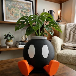 May include: A spherical black planter shaped like a Bob-omb character with two large white oval eyes and two bright orange feet. The planter has a grey rim at the top and contains a green Monstera adansonii plant with several perforated leaves.