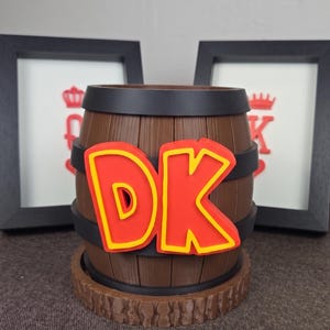 May include: A brown barrel-shaped container with the letters "DK" in red with yellow outlines. The container has black bands and sits on a wooden base. Two framed pictures are in the background.