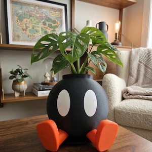 May include: A black, round planter with white oval eyes and orange feet, resembling a cartoon bomb. The planter holds a green plant with large, hole-filled leaves. The planter sits on a wooden table in a well-lit room.