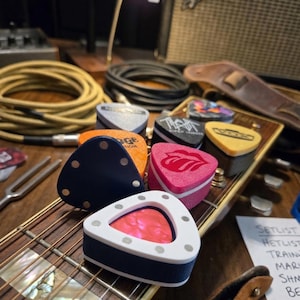 May include: A collection of guitar picks in various colors and designs, including a navy blue pick with silver dots, a pink pick with a red lip design, and a white and blue pick with a red interior. The picks are arranged on a guitar.