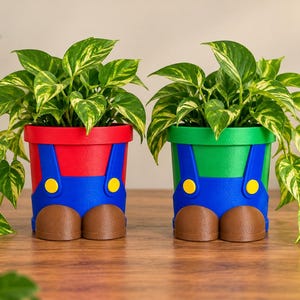 May include: Two cylindrical plant pots featuring a design of blue overalls with yellow buttons and brown shoes. The left pot has a red upper section, and the right pot has a green upper section. Each pot contains a lush, variegated green pothos plant.