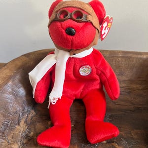 Puede incluir: Oso de peluche rojo Beanie Baby con un gorro de aviador marrón con gafas y una bufanda blanca. El oso tiene una etiqueta Ty pegada a la oreja y un parche de 100 Years of Flight en el pecho.