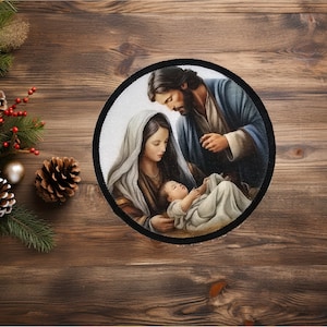 May include: A round, framed image of a traditional depiction of the Holy Family. Mary, Joseph, and baby Jesus are depicted in a loving embrace. The image is set against a white background and framed with a black border.