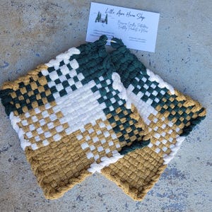 May include: Two square, woven potholders in shades of gold, white, and dark green. The potholders have a checkerboard pattern and a loop for hanging. A small card with text is in the background.