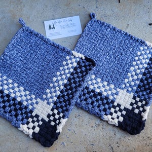 May include: Two woven potholders in shades of blue, white, and black. The potholders have a square shape with a loop for hanging. The design features a woven pattern with a color block design. The potholders are handmade.