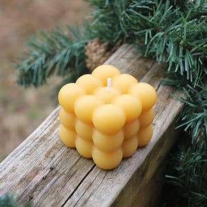 Puede incluir: Una vela cuadrada y amarilla con burbujas y una mecha blanca, apoyada sobre una superficie de madera envejecida. La vela está rodeada por un fondo borroso de agujas y ramas de pino verdes, creando una estética natural y rústica.