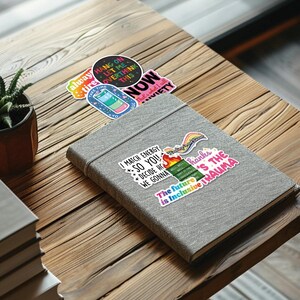 May include: A gray notebook with colorful stickers on the cover. The stickers feature phrases like "I match energy so you decide how we gonna" and "The future is inclusive trauma".